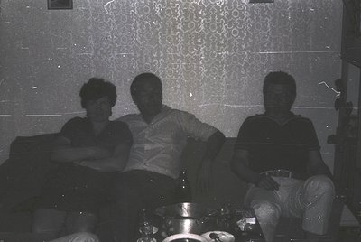 Three individuals pose indoors on a patterned sofa, likely mid-20th century (1960s–1970s). The woman wears a sleeveless dress...