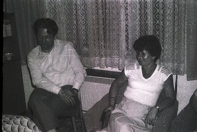 Two individuals seated indoors in mid-20th century attire, likely 1960s–1970s. Man in button-down shirt, woman in patterned b...