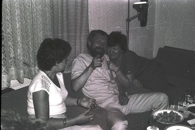 Vintage indoor gathering featuring three adults in casual 1970s attire—man in a light-colored button-up shirt, woman on left ...