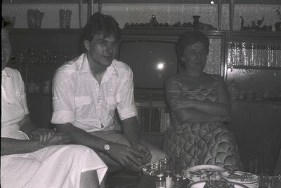 Black-and-white interior shot of three individuals seated at a table in a dimly lit room, likely a 1970s café or restaurant. ...