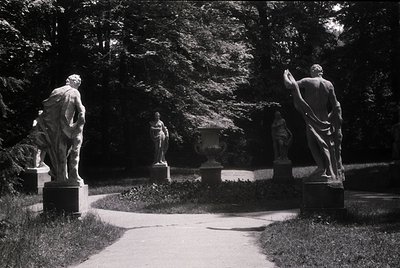 Neoclassical stone statues flanking a sunlit pathway in a lush garden, likely 19th–early 20th century. Four figures—two male ...
