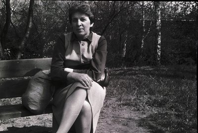 Black-and-white portrait of a woman seated on a wooden bench in a forested area, mid-20th century. She wears a structured bla...