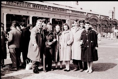 Mid-20th century group photo outside grand neoclassical train station. Seven individuals in 1950s attire—men in suits/coats, ...