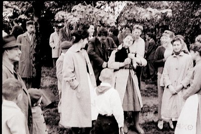 Vintage black-and-white outdoor gathering, likely mid-20th century. Crowd in autumn foliage, featuring men in suits, women in...