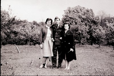 Three individuals pose outdoors in mid-20th-century attire, likely 1940s–1950s. The man wears a military uniform with insigni...