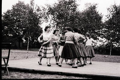 Group of women in 1950s-style dresses and pleated skirts performing outdoors on a wooden platform, likely a folk or dance ens...