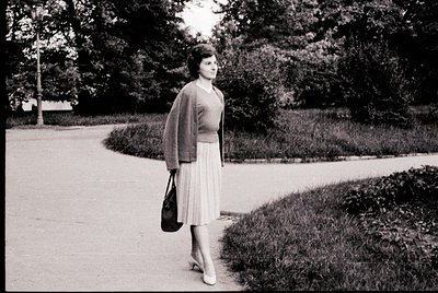 Mid-century woman in a fitted cardigan and pleated skirt strolls along a tree-lined park path, holding a structured handbag. ...