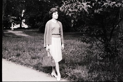 Mid-century woman in a knee-length skirt and cardigan strolls along a tree-lined park path, holding a structured handbag. Sof...