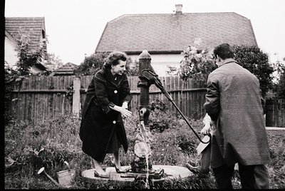 Mid-20th century (likely 1950s–1960s) black-and-white photo showing two individuals operating a manual well pump in a residen...