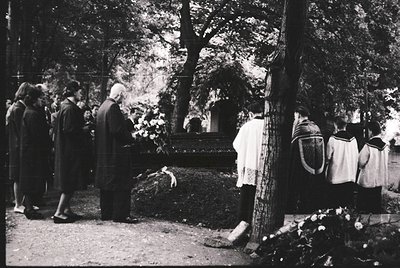 Black-and-white memorial scene in a forested cemetery, mid-20th century. Group of mourners, including a man in suit and women...