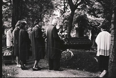 Mid-20th century funeral procession in a wooded cemetery. Group of mourners in formal attire—men in suits, women in long dres...