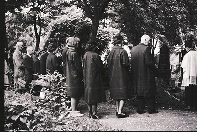 Black-and-white group standing at graveside in wooded cemetery, mid-20th century. Men in suits, women in long coats and hats,...