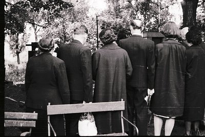 Group of six individuals in mid-20th century attire stands at a gravesite, facing crosses. Men wear suits, women in dresses w...