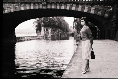A woman in mid-20th-century fashion stands under a stone bridge arch beside a canal, holding a small handbag. Her skirt, card...
