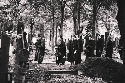 Black-and-white cemetery scene with seven mourners in traditional Eastern European attire (long coats, headscarves) gathered ...