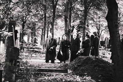 Mid-20th century black-and-white cemetery scene with overgrown graves and dense forest backdrop. Four mourners in dark clothi...