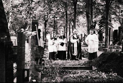 Mid-20th century black-and-white cemetery scene: clergy in liturgical vestments (cassocks, stoles) conduct a funeral service ...