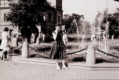 Vintage black-and-white photo of a woman in a 1950s-style dress with floral appliqué, posing near a fountain in an urban park...
