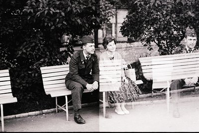 Vintage black-and-white photo of two individuals seated on park benches in a shaded area, likely mid-20th century. The man we...