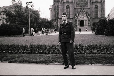 Mid-20th century uniformed man in a landscaped urban park, Gothic-style church with pointed arches in background. Black-and-w...