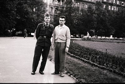 Black-and-white portrait of two men in 1950s European urban park. Left: uniformed individual in peaked cap, belted jacket, an...