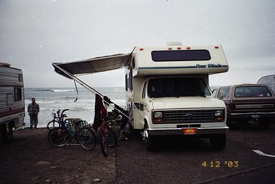 Classic 1980s-era Four Winds RV parked by a coastal road, with extended awning. Two bicycles secured to the front, one with a...