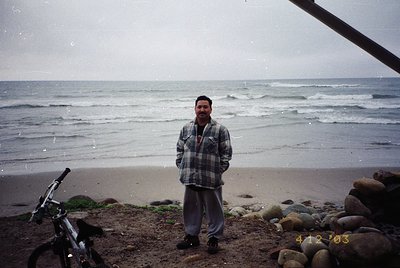 Man in plaid shirt and dark pants stands on a rocky seaside beach, rain-soaked waves crashing behind. Black bicycle leans aga...