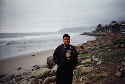 Young man in a black jacket with "AMER" embroidered on the back poses on a rocky beach during light snowfall, with coastal cl...