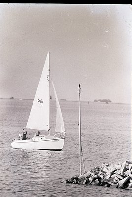 Vintage monochrome sailboat race photo, 1960s-70s. Small fiberglass dinghy () with two crew members, sailing near a rocky bre...