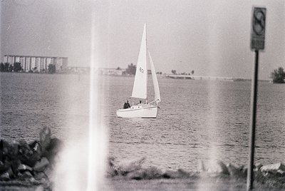 Vintage black-and-white sailboat on calm waters, framed by window or glass. Mid-20th century coastal architecture in backgrou...