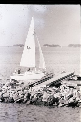 Vintage monochrome photo of a small sailboat () grounded on rocky shore, mid-20th century. Two figures aboard; distant landfo...