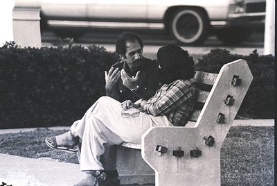 Two men in mid-20th-century attire share a moment on a modernist concrete bench, likely 1960s–1970s. The man on left wears li...