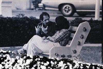 Black-and-white candid of two individuals seated on a modernist concrete bench in a landscaped park setting, 1960s–1970s. One...
