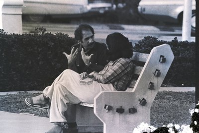 Two men in a candid 1970s-era conversation on a modernist white bench in an outdoor setting. The seated man wears light-color...