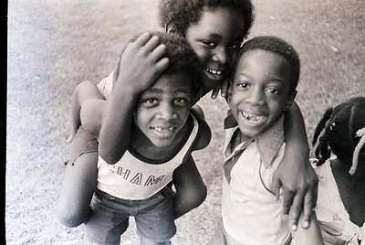 Black-and-white candid of three joyful children posing outdoors, likely mid-20th century. The boy in the center wears a "CHAM...