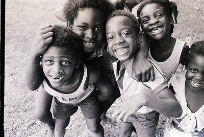Four joyful children pose closely in a 1970s outdoor setting, wearing matching white tank tops with floral patterns. Their br...