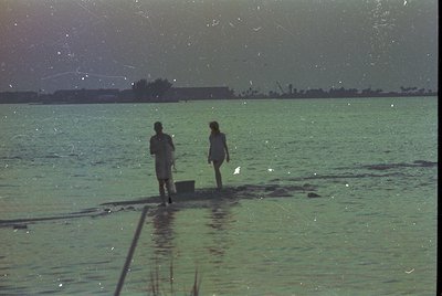 Vintage seaside scene featuring two figures walking on shallow water near a dock, likely mid-20th century. Green-tinted film ...