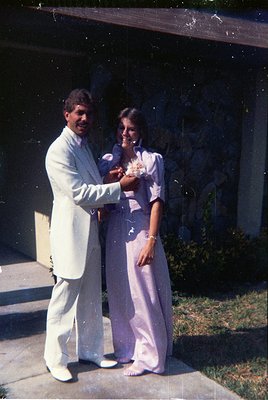 1970s formal portrait of a man in a white suit and woman in a pastel pink gown, holding a bouquet. Outdoor setting with blurr...