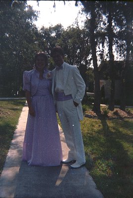 Couple in formal attire—woman in a fitted purple gown with a high collar, man in a light-colored suit with a sash—posing outd...