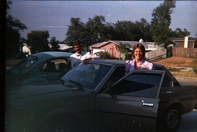 Vintage 1970s suburban scene: two people pose in a dark sedan (likely a Volkswagen Passat) with a man in a suit and woman in ...