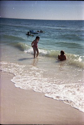 Vintage seaside scene with two children wading in shallow waves, one standing in a striped shirt, the other kneeling in swimw...