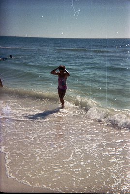 Vintage seaside scene: Woman in 1960s-style one-piece swimsuit wading into shallow waves, hand shielding eyes from sun. Light...