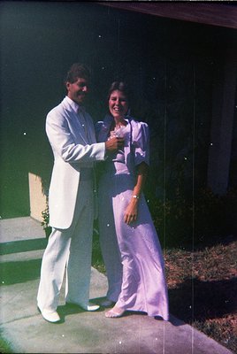 Two individuals pose indoors, likely at a 1970s wedding or formal event. The man wears a white suit with a satin lapel, while...