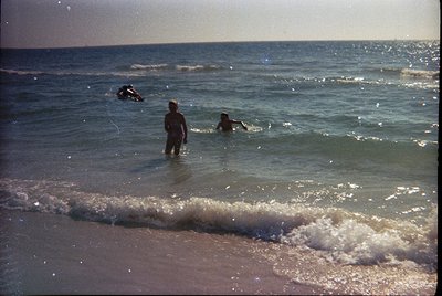 Vintage seaside scene: three men wading in shallow waves, one standing near shore, two swimming offshore. Gentle surf meets s...