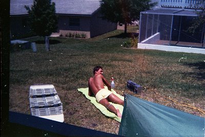 Vintage backyard scene featuring a shirtless man reclining on a green towel, holding a plastic bottle. Surrounding elements i...