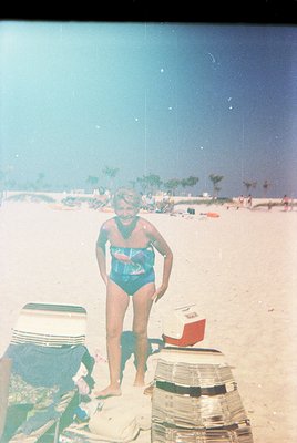 Vintage seaside photo featuring a woman in a teal one-piece swimsuit, standing on sandy beach with a red cooler and striped t...