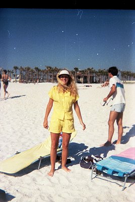 Vintage beach scene with woman in bright yellow 1970s-style swimsuit and straw hat, posing on white sand. Palm trees and beac...