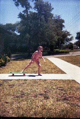 Vintage photo of a child skateboarding on a residential street, 1970s–1980s. Red-and-white striped shirt, barefoot, on a gree...