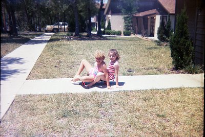Two young girls pose on a suburban sidewalk in the 1970s, wearing patterned swimsuits. Grass, trees, and a brick driveway fra...