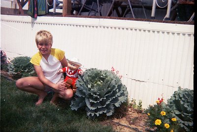 Child kneeling beside large cabbage plants in a garden, holding a toy. Mid-20th century suburban backyard with white picket f...